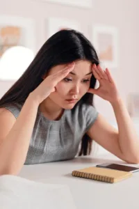 Asian woman experiencing stress, sitting at desk with hands on her head, indoors.